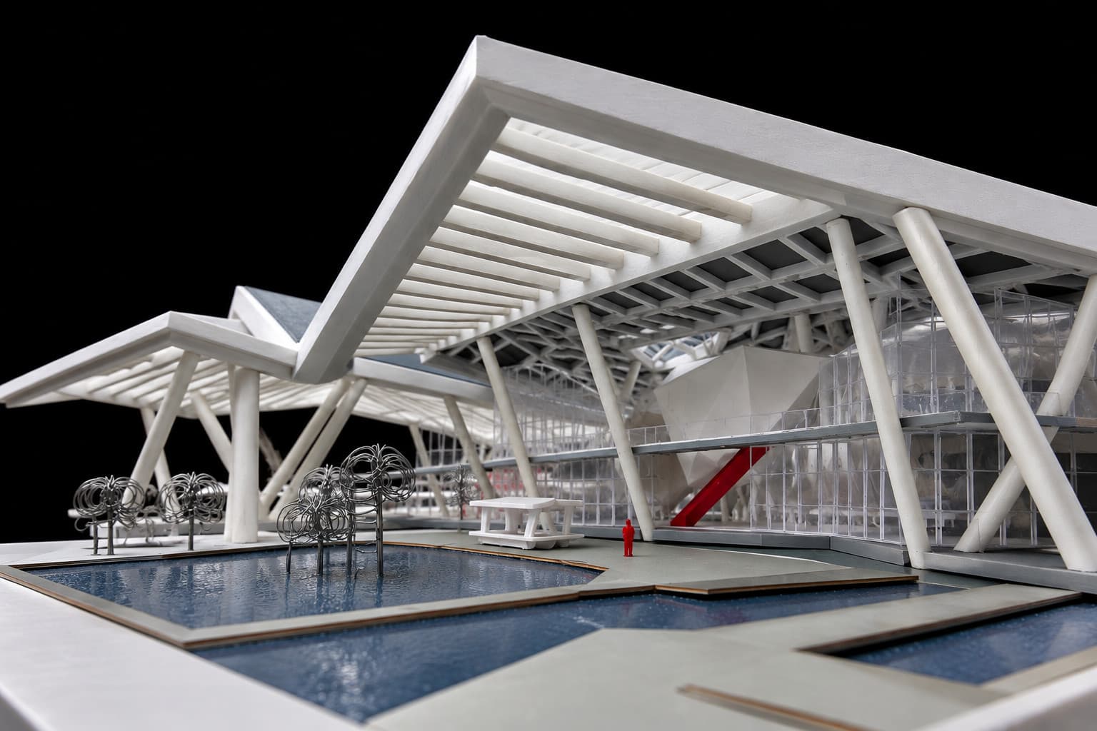 A white 3D printed scale model of a public building with a folded roof on slender columns, glazing and pools of water
